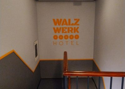 Treppenhaus mit orangefarbenem Schriftzug 'WALZWERK HOTEL' an der Wand.